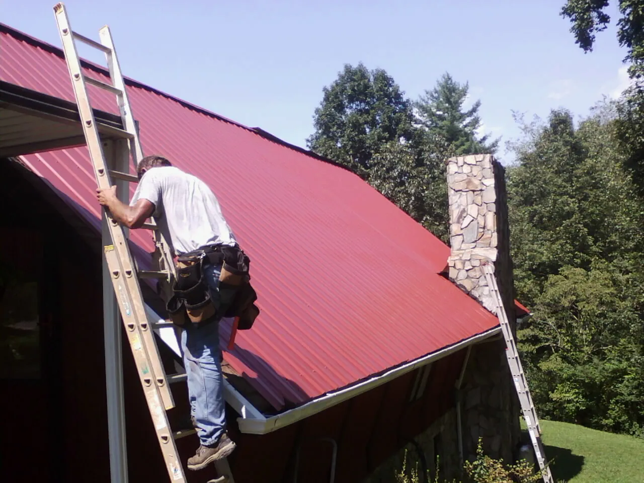 Roofer on a ladder beside a red metal roof and stone chimney — Soffit & Fascia Repair by Roofing Brothers in Rio Linda